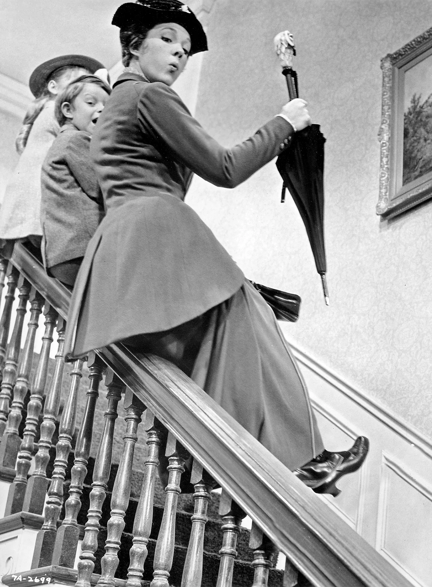 Julie Andrews, playing Mary Poppins, slides down the bannister of a staircase with two children, Jane and Michael, in tow.